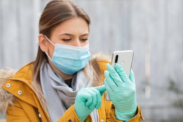 Portrait young woman with protective mask and gloves holding smartphone - coronavirus pandemic alert - Photo, image
