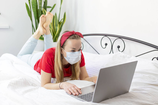 mujer joven enferma con una máscara médica y una camiseta roja y pantalones cómodos se encuentra en una cama blanca con un ordenador portátil. Trabaja desde casa debido a la epidemia de coronavirus. pandemia COVID-19
 - Foto, Imagen