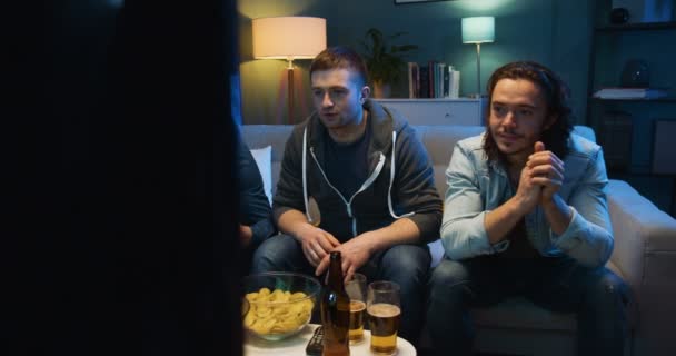 Caucasian happy friendly guys watching TV on couch at home in evening and cheering for favorite sport team in football match. Male fans resting at night in front of television screen. - Materiał filmowy, wideo