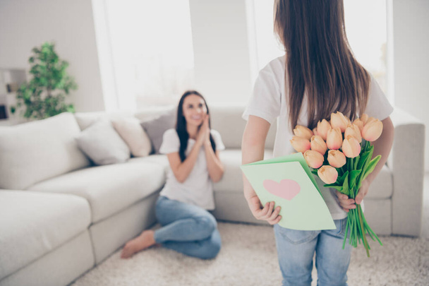 Photo of pretty funny little girl hiding behind back young charming brunette mommy birthday spring present postcard tulips flowers positive affection emotions home indoors - Fotografie, Obrázek