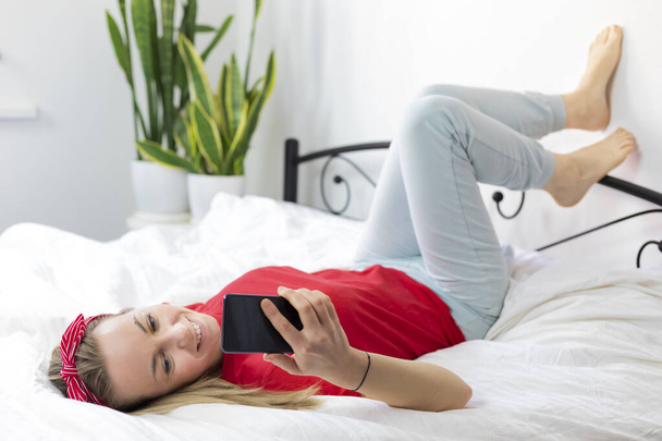 young smiling woman with blond hair in a red T-shirt and comfortable pants lies on a white bed with a smartphone. she works from home, writes and reads messages, lounges - Photo, Image