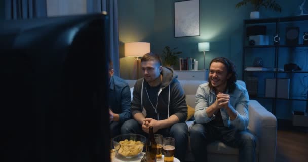 Male football fans sitting on couch in front of TV screen and cheering for favorite team with beer and snacks at night. Guys celebrating win in match and hugs while spending time together in evening. - Filmagem, Vídeo