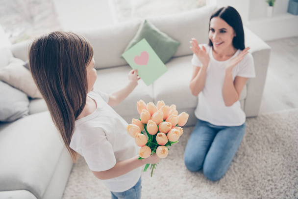Photo of pretty funny little girl giving young charming brunette mommy birthday spring present postcard fresh tulips flowers positive affection emotions home indoors - Fotó, kép