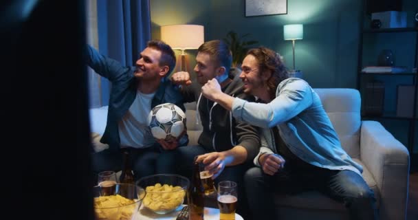 Three cheerful Caucasian friendly men sitting on sofa at night with football ball, snacks and beer and taking selfie photo with smartphone camera. Sport fans posing to phone in living room at TV. - Video, Çekim