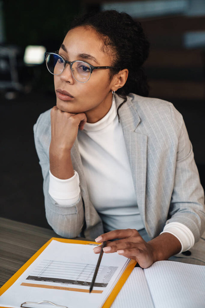 Image of young brunette african american businesswoman sitting at table and working with papers in office room - Photo, Image