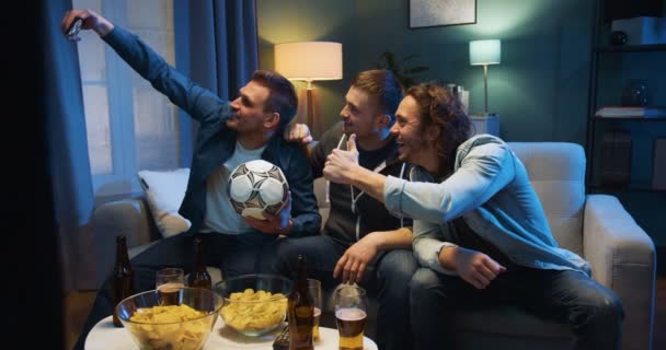 Three handsome Caucasian guys best friends sitting on couch in evening with football ball, snacks and beer and taking selfie photo with smartphone camera. Sport fans posing to phone at home at TV. - Filmati, video