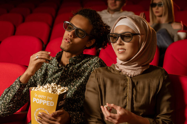 African american muslim man with his wife sitting in movie theater, watching 3D movie, eating popcorn, smiling. - Foto, Bild