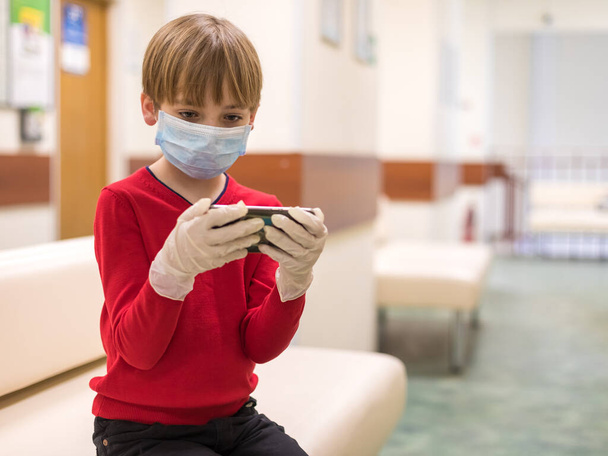 Caucasian boy in a red sweater with a medical mask on his face plays in the phone. The child 's hands are wearing gloves. The concept of visiting public places during the disease epidemic - Fotó, kép