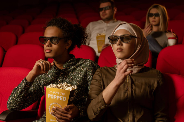 Африканский американец-мусульманин с женой, сидящей в кинотеатре, смотрящей 3D фильм, кушающей попкорн, улыбающейся
. - Фото, изображение