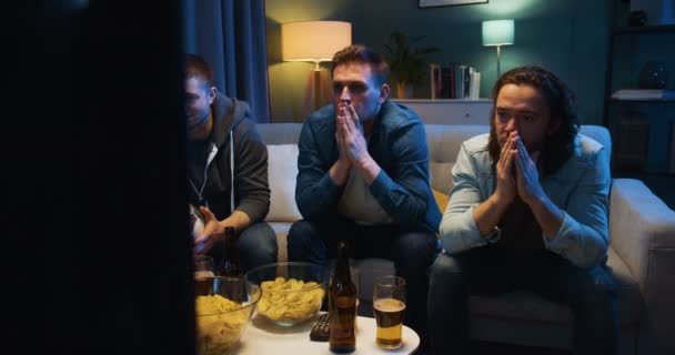Three Caucasiand sad and worried guys friends sitting on sofa at home in dark living room and watching football match. Favourite team loosing concept. Dissapointed men in front of sport channel. - Materiał filmowy, wideo