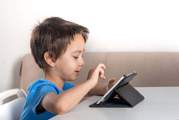 Tiro de menino divertido sentado em uma mesa em casa e navegando no internrt em um tablet. Estilo de vida moderno
.   - Foto, Imagem