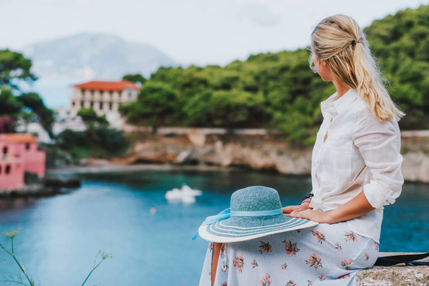 Beautiful blond woman holding blue sunhat sitting and enjoying blue bay of colorful tranquil village Assos, Kefalonia, Greece - Photo, image