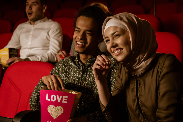 African american muslim man with his wife sitting in movie theater, watching movie, eating popcorn, smiling. - Photo, Image