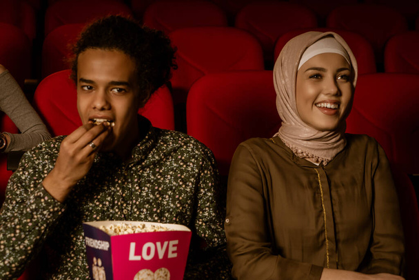 African american muslim man with his wife sitting in movie theater, watching movie, eating popcorn, smiling. - Foto, Bild