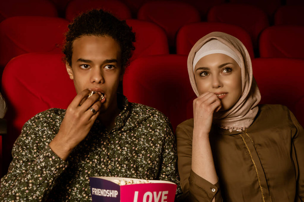 African american muslim man with his wife sitting in movie theater, watching movie, eating popcorn, smiling. - Photo, image