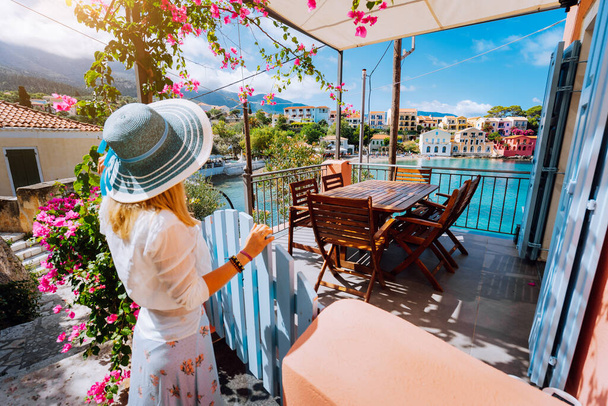 Female tourist with blue sun hat staying in Assos village in front of cozy veranda and enjoying turquoise colored bay of Mediterranean sea and beautiful colorful houses of in Kefalonia, Greece - Foto, Bild