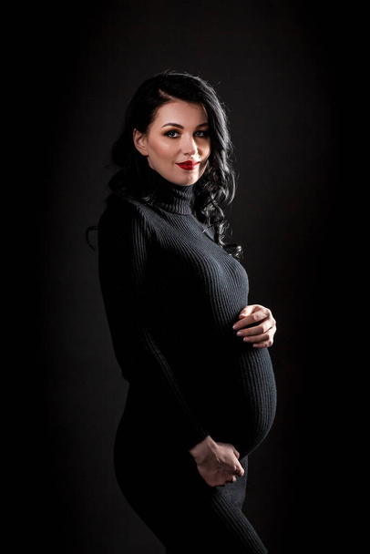 Mladá krásná štíhlá těhotná žena s dlouhými vlasy v černém těle a boty stojící a objímající břicho přes tmavé pozadí. Těhotenství módní vzhled koncept. - Fotografie, Obrázek