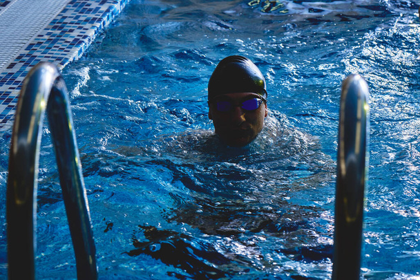 Close-up portrait of a sporty muscular man in swimming goggles and a cap in a swimming pool indoors - Photo, Image