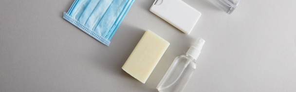 Sprey şişelerdeki el dezenfektanı, tıbbi maske ve gri arka planda antibakteriyel sabun, panoramik çekim - Fotoğraf, Görsel