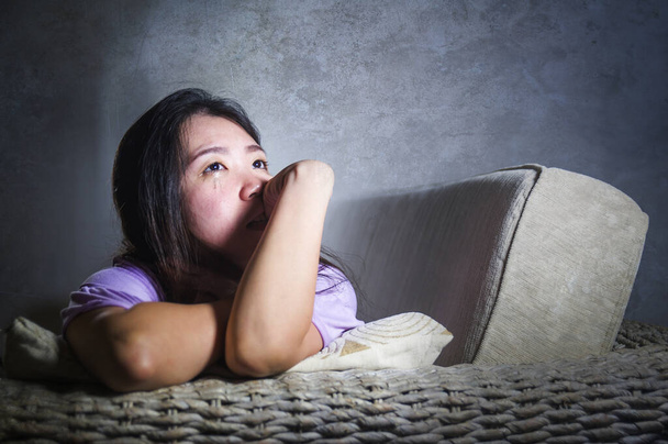 covid-19 virus home lockdown - dramatic portrait of young beautiful depressed and scared Asian Chinese woman lying sad and desperate on sofa couch feeling lonely and in panic about world crisis  - 写真・画像