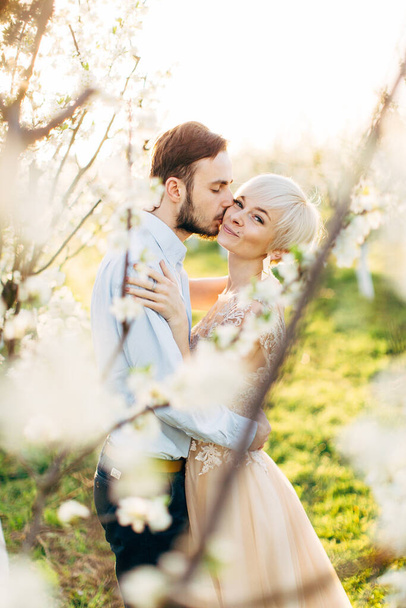 Happy couple in elegant luxury wear, walking in beautiful blooming garden, enjoying spring nature. Pretty woman looking at camera and smiling while her handsome man kissing her cheek - Foto, imagen