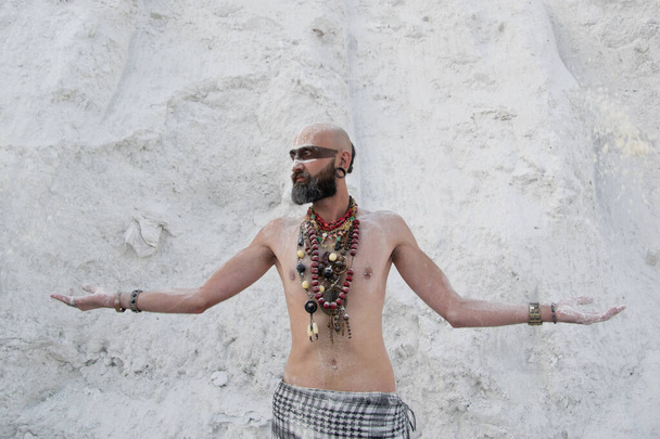 νεαρός άνδρας με πρωτόγονο μακιγιάζ και χάντρες ξόρκια με σκόνη στα χέρια σε λευκά βράχια - Φωτογραφία, εικόνα