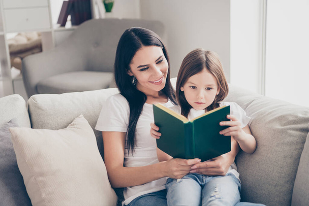 Photo of pretty little girl young charming mommy having fun reading interesting book sitting comfortable sofa spend free time weekend together indoors home house - Фото, зображення
