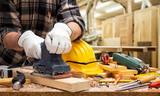 Close-up. O carpinteiro com as mãos protegidas por luvas com a lixadeira elétrica alisa uma tábua de madeira. Indústria da construção, oficina de carpintaria
. - Foto, Imagem