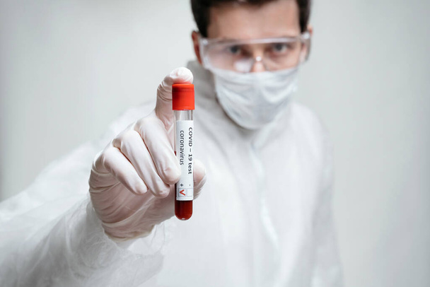 Scientist in protective suit, mask and gloves is holding positive COVID-19 test with sample of blood in laboratory tube on white background. Pandemic infectious concept - Фото, изображение