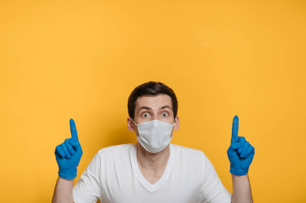 Scared young man in protective face mask and medical gloves pointing to empty space on background. Copy space, isolated on yellow background. Panic and home isolation due to coronavirus outbreak. - Photo, Image