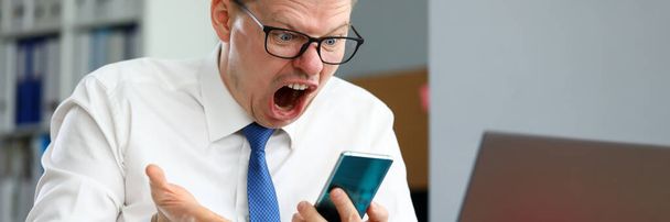 Hombre de negocios infeliz sentado en la mesa de trabajo gritando en el teléfono móvil
 - Foto, Imagen