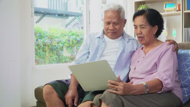 Gelukkig Aziatisch echtpaar zit op de bank en met behulp van een laptop om te surfen op het internet en VIDEO conferentie met familie. Begrip technologie brengt gezinnen dichter bij elkaar, sociale afstand - Video