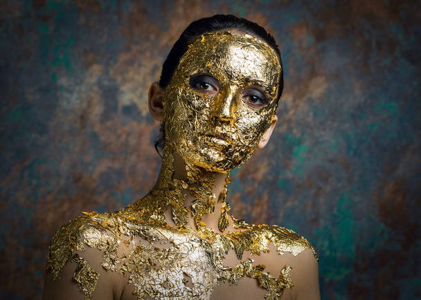 Tyttö, jolla on kultaisesta lehdestä tehty naamio kasvoillaan. Gloomy studio muotokuva brunette abstrakti tausta. - Valokuva, kuva