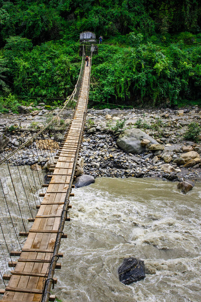 Narrow hanging bridge over mountain river in Himalayas, Nepal, with a person standing at the far end of the bridge. - Photo, Image