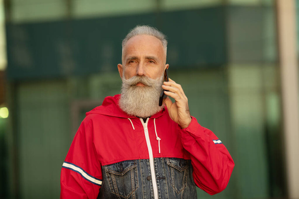 Handsome senior bearded man is talking by a smartphone while having coffee break on a street beside office building. - Фото, изображение