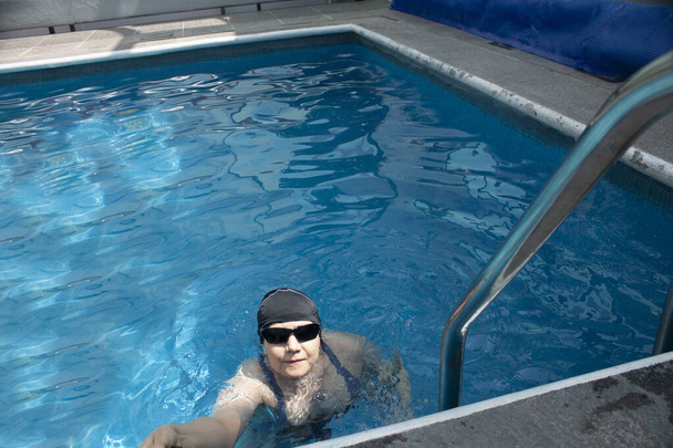 Donna con la capacità di galleggiare in una fossa ed estendere il braccio per raggiungere l'oggetto fuori dalla piscina mentre muove le gambe per rimanere in piedi in acqua tra onde e bolle indossa un cappello e occhiali da sole per la protezione solare
 - Foto, immagini