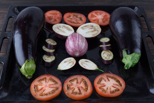 Whole and sliced eggplants and tomatoes on rustic baking tray. Shallow depth of field - Foto, Imagen