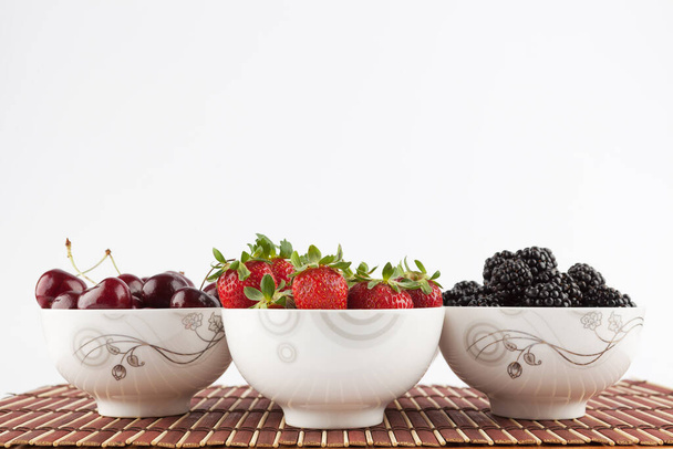 Three ceramic bowls with berries - Strawberries, Blackberries, and Cherries on bamboo mat isolated on white - Photo, image