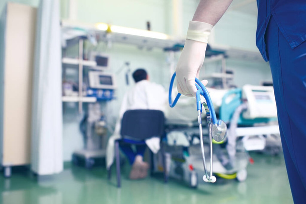 Figur eines Arztes mit Stethoskop in den Händen auf dem Hintergrund eines Mannes, der in der Nähe des Patientenbettes auf der Krankenstation sitzt. - Foto, Bild