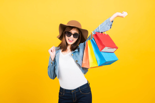 Asian happy portrait beautiful cute young woman teen smiling standing with sunglasses excited holding shopping bags multi color looking camera isolated, studio shot yellow background with copy space - 写真・画像