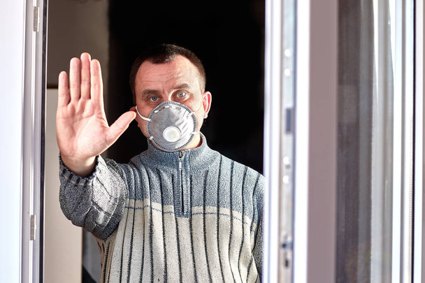 Een man met een medisch masker op zijn gezicht staat achter een witte deur en laat ons de halte zien. Close-up. - Foto, afbeelding