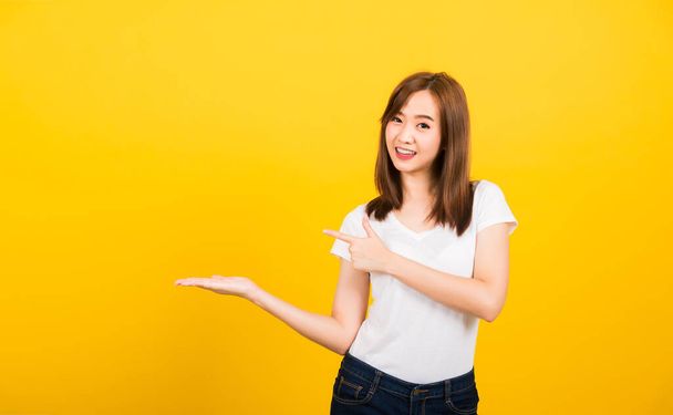 Asian happy portrait beautiful cute young woman teen standing wear t-shirt holding something on palm and point away side looking to camera isolated, studio shot on yellow background with copy space - Photo, Image