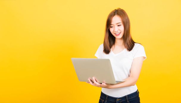 Asian happy portrait beautiful cute young woman teen smiling standing wear t-shirt using laptop computer on hand looking to computer isolated, studio shot on yellow background with copy space - Photo, Image