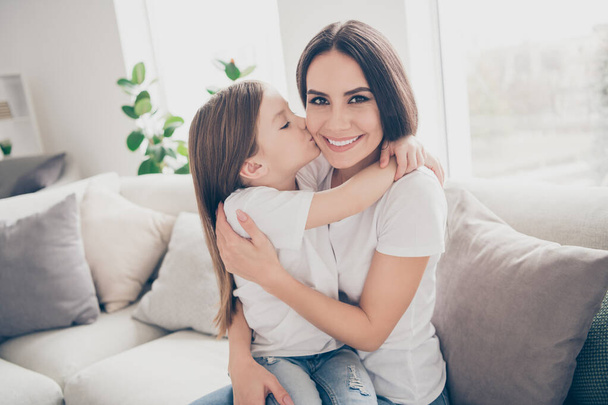 Portrait of positive cheerful sweet mommy and her little girl kiss mommy cuddle embrace enjoy spend free time together sit divan in light room apartment indoors - Foto, Imagem