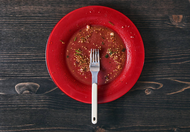 Salad leftovers after meal. Oil, tomato seeds, chopped parsley on a single red plate with white fork. Wooden table background. Top down flat photo with copy space - Photo, image