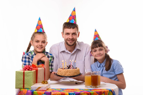 Ο μπαμπάς και δύο κόρες με μια τούρτα στο τραπέζι με χαρά κοιτάζουν το πλαίσιο. - Φωτογραφία, εικόνα