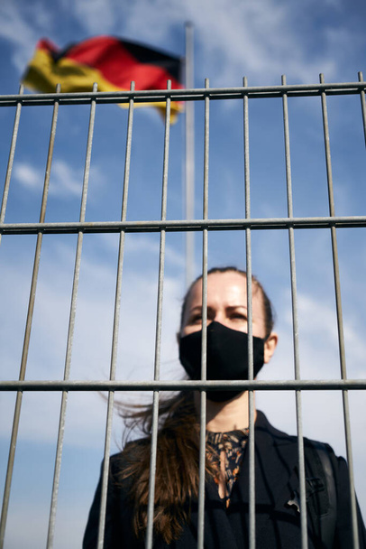 Menina loira de pele clara com uma máscara protetora preta em seu rosto, fica atrás da grade de bloqueio, a bandeira balança no fundo
 - Foto, Imagem