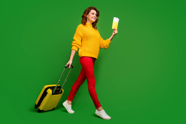 Full size profile photo of cheerful lady hold tickets passport travel abroad rolling case go flight registration table wear yellow sweater red pants shoes isolated green color background - Фото, изображение