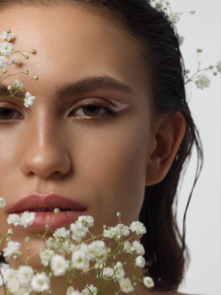 Close up portrait of a face with beautiful skin and nude makeup, eyeliner and long eyelashes, puffy natural lips and white flowers. Fashion photography. Cosmetology and spa. Freshness of spring - 写真・画像
