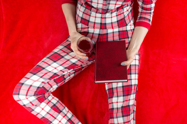 a woman in red checked home suite with a book and glass of wine at home on a red background in quarantine coronavirus covid-19, without face - Valokuva, kuva